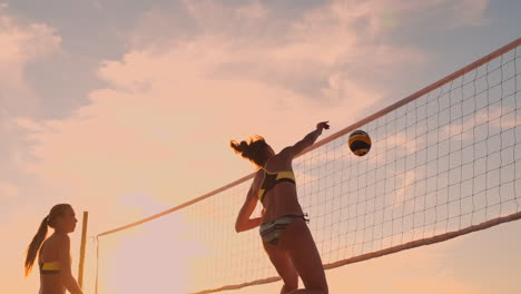 Cámara-Lenta,-ángulo-Bajo,-Primer-Plano,-Destello-De-Lente:-Jóvenes-Jugadoras-De-Voleibol-Pasan-Y-Rematan-La-Pelota-Sobre-La-Red-En-Una-Soleada-Tarde-De-Verano.-Chicas-Caucásicas-En-Forma-Jugando-Voleibol-De-Playa-Al-Atardecer.